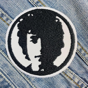 May include: A round, black and white embroidered patch featuring a silhouette of a person's face. The black silhouette is set against a white background, with a white border. The patch is placed on a denim jacket.