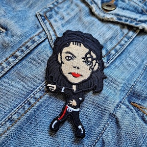 May include: Embroidered patch featuring a cartoon depiction of a pop icon in a black outfit with red accents. The patch has a black border and is set against a blue denim jacket.