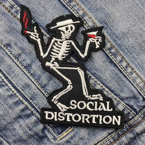May include: Black and white embroidered patch featuring a skeleton figure holding a lit cigarette and a drink. The skeleton wears a hat and is set against a black background. The words "SOCIAL DISTORTION" are displayed below the skeleton.