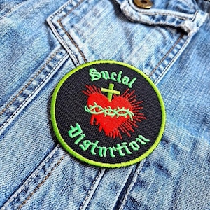 Può includere: Una toppa rotonda nera con bordi verdi raffigurante un cuore rosso trafitto da una corona di spine e una croce. Le parole "Social Distortion" sono ricamate in verde attorno al cuore.