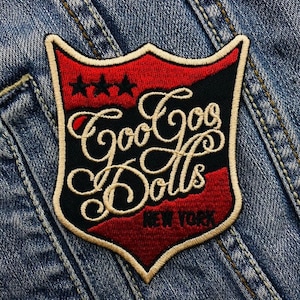 May include: An embroidered patch in red, white and black with the text "Goo Goo Dolls" and "New York" in a script font. The patch is shaped like a shield with three stars at the top.