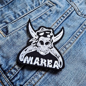 Könnte beinhalten: Schwarz-weißer, gestickter Aufnäher mit einem Totenkopf, Hörnern und dem Wort "MAREA". Der Aufnäher ist auf eine blaue Jeansjacke genäht. Der Aufnäher hat ein detailliertes Design mit weißen Nähten.