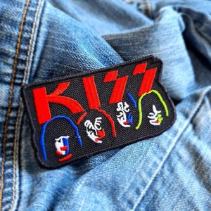 May include: Black and red embroidered patch with the word "KISS" in red letters and four cartoon faces in blue, red, green, and yellow.