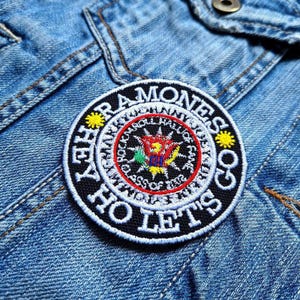 May include: A black and white embroidered patch with the words "The Ramones Let's Go" and a red, yellow, green and blue starburst design in the centre. The patch is on a blue denim jacket.