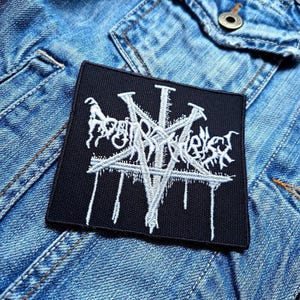 May include: Black embroidered patch with a white design of a pentagram with a stylized text "Vomit The Soul" inside. The patch is on a blue denim jacket.