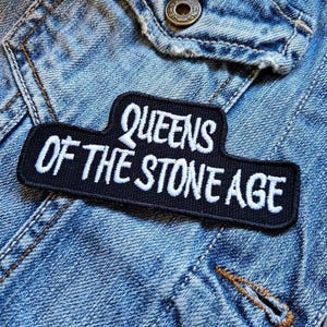 Könnte beinhalten: Ein gestickter schwarzer und weißer Aufnäher mit dem Text "Queens of the Stone Age" auf einer Jeansjacke.