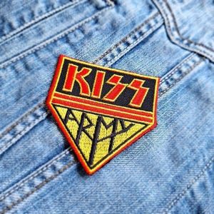May include: A black, red, and yellow embroidered patch with the word "KISS" in red and black letters and the word "ARMY" in black letters on a yellow background. The patch is in the shape of a diamond.