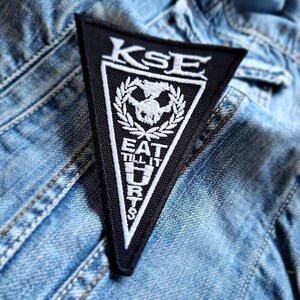 May include: Black triangular patch with white text and a skull design. The patch reads "KSE" at the top, and "EAT TILL IT HURTS" at the bottom. The patch is on a denim jacket.