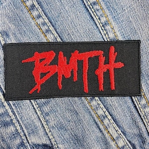 Puede incluir: Un parche rectangular negro con las letras rojas "BMTH" bordadas. El parche está cosido en una chaqueta vaquera. Las letras están en una fuente estilizada y en negrita.