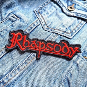 May include: A black and red embroidered patch with the word "Rhapsody" in a stylized font. The patch is sewn onto a blue denim jacket.