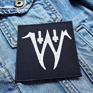 Puede incluir: Parche de tela negro con un logotipo bordado en blanco de una "W" estilizada con dos cruces a cada lado.