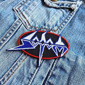 Può includere: Una toppa ricamata in bianco e nero con un contorno rosso. La toppa presenta un logo stilizzato con la parola "SLAYER" in un font frastagliato.