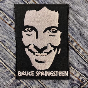 May include: A black embroidered patch featuring a stylized portrait of Bruce Springsteen in white thread. The text "BRUCE SPRINGSTEEN" is embroidered in white below the portrait. The patch is rectangular and set against a denim background.