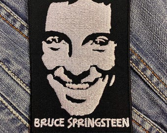 May include: A black embroidered patch featuring a stylized portrait of Bruce Springsteen in white thread. The text "BRUCE SPRINGSTEEN" is embroidered in white below the portrait. The patch is rectangular and set against a denim background.