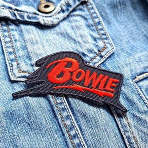 May include: A black and red embroidered patch with the word "BOWIE" in red lettering on a denim jacket.