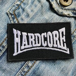 Può includere: Una toppa rettangolare nera con la scritta "HARDCORE" ricamata in bianco, con un lettering stilizzato. La toppa è posta su uno sfondo di giacca di jeans, suggerendo il suo utilizzo su abbigliamento o accessori.