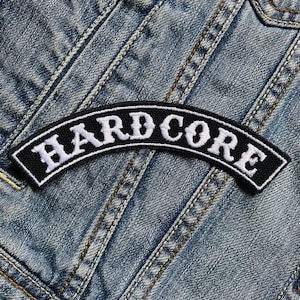 Könnte beinhalten: Schwarz-weißer gestickter Aufnäher mit dem Wort "HARDCORE" in weißen Buchstaben auf schwarzem Hintergrund. Der Aufnäher ist auf eine blaue Jeansjacke genäht.