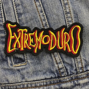 Puede incluir: Un parche bordado negro con la palabra "EXTREMODURO" en letras amarillas y rojas. El parche está cosido en una chaqueta vaquera. Las letras tienen una apariencia estilizada y manuscrita.