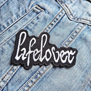 May include: A black and white embroidered patch with the word "lifelover" in a stylized font. The patch is sewn onto a blue denim jacket.