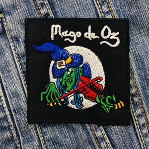 Könnte beinhalten: Schwarzer gestickter Patch mit einer grünen Hexe, die eine rote Geige spielt. Der Text "Mago de Oz" ist in Weiß über der Hexe gestickt.