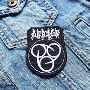 May include: Black and white embroidered patch with the text "DECADE" and a stylized symbol of three interlocking circles. The patch is sewn onto a blue denim jacket.