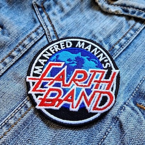 Könnte beinhalten: Ein runder Aufnäher mit dem Text "MANFRED MANN'S EARTH BAND" in roter und weißer Schrift. Der Aufnäher hat ein blau-weißes Globus-Design in der Mitte, vor einem schwarzen Hintergrund. Der Aufnäher ist auf eine Jeansjacke genäht.