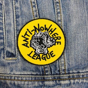 Puede incluir: Parche bordado amarillo con texto negro que dice "Anti Nowhere League" y un gráfico en blanco y negro de una criatura que gruñe.