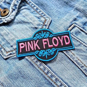 Op de afbeelding: Een blauwe en zwarte geborduurde patch met de tekst "Pink Floyd" in roze letters.