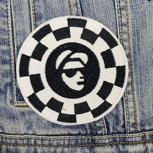 Könnte beinhalten: Runder, schwarz-weißer Aufnäher mit Schachbrettmuster-Rand. In der Mitte befindet sich eine stilisierte schwarze Silhouette einer Person mit Sonnenbrille und Hut. Der Aufnäher ist auf eine Jeansjacke genäht.