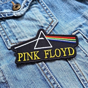 Könnte beinhalten: Schwarz-weiß gestickter Patch mit einem Dreieck und einem Regenbogen, mit dem Text "PINK FLOYD" in gelben Buchstaben. Der Patch ist auf eine blaue Jeansjacke genäht.