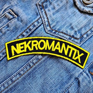 Pode incluir: Um remendo bordado preto e amarelo com a palavra "NEKROMANTIX" em letras amarelas.