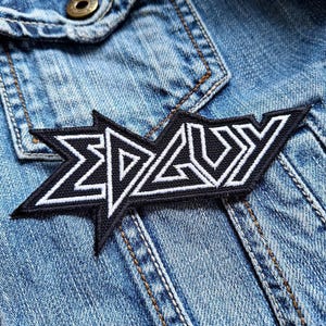 Op de afbeelding: Zwart-wit geborduurde patch met het woord "EDGUY" in een gestileerd lettertype. De patch is bevestigd aan een blauw denim jack.