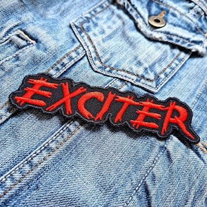May include: A black and red embroidered patch with the word "EXCITER" in a bold, stylised font. The patch is sewn onto a blue denim jacket.