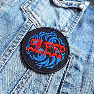 Pode incluir: Um remendo bordado preto e azul com a palavra "SLEEP" em letras vermelhas. O remendo é circular e tem um design de ondas.