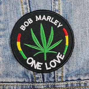 Può includere: Toppa ricamata nera e verde con il testo "Bob Marley One Love" e una foglia di marijuana stilizzata. Il testo è bianco e lo sfondo è nero. La toppa ha un bordo rosso, giallo e verde.