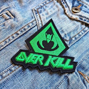 Pode incluir: Um patch bordado preto e verde com o texto "OVER KILL". O patch apresenta um design gráfico verde de uma figura com chifres dentro de um losango. O patch é costurado em uma jaqueta jeans.