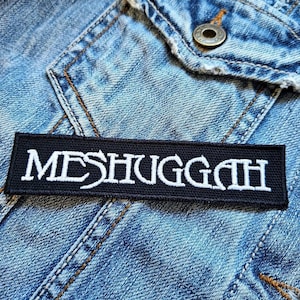 Può includere: Toppa ricamata in bianco e nero con la parola "MESHUGGAH" in lettere bianche su sfondo nero.