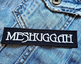 Może przedstawiać: Czarno-biała naszywka haftowana z napisem "MESHUGGAH" w białych literach na czarnym tle.