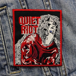 May include: A black and red embroidered patch featuring the words "Quiet Riot" and a white image of a person wearing a mask.