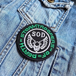May include: A black and green embroidered patch with the text "Stormtroopers of Death" and "SOD" in a white circle with a white skull and wings inside.