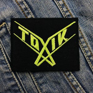 Pode incluir: Um remendo preto com um logotipo bordado verde que diz "TOXIK" em uma fonte estilizada.