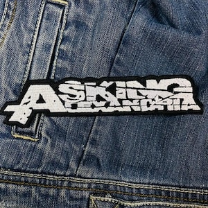 Könnte beinhalten: Ein rechteckiger Aufnäher mit dem Text "ASKING ALEXANDRIA" in weißer, stilisierter Schrift, umrandet von Schwarz. Der Aufnäher ist auf einer Jeansjacke angebracht, mit sichtbaren Nähten.