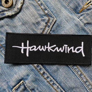 May include: A black rectangular patch with the white embroidered text "Hawkwind". The patch is set against a blue denim jacket, with visible seams and a button.