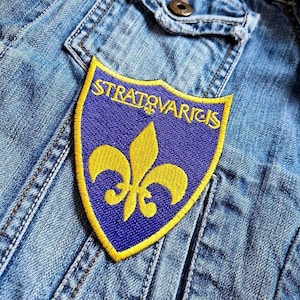 Puede incluir: Parche bordado azul y amarillo con el texto "STRATOVARICS" y un diseño de flor de lis. El parche está cosido en una chaqueta vaquera.