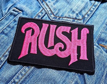 Könnte beinhalten: Ein schwarzer Stoff-Patch mit dem Wort "RUSH" in pinker Stickerei. Der Patch ist auf eine blaue Jeansjacke genäht.