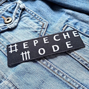 Op de afbeelding: Zwarte patch met witte tekst die "#Depeche Mode" zegt. De patch zit op een blauw denim jack.