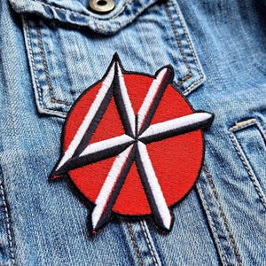 May include: Red and white embroidered patch with a starburst design. The patch is sewn onto a blue denim jacket.