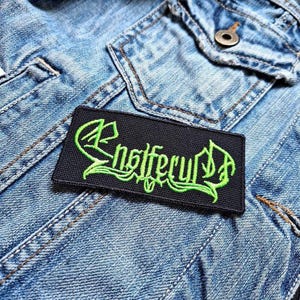 Op de afbeelding: Zwarte patch met de bandnaam "Enslaved" geborduurd in groen.