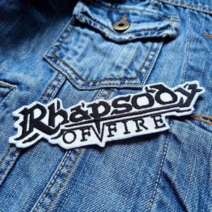 Può includere: Una toppa ricamata in bianco e nero con il testo "Rhapsody Of Fire" su una giacca di jeans blu.