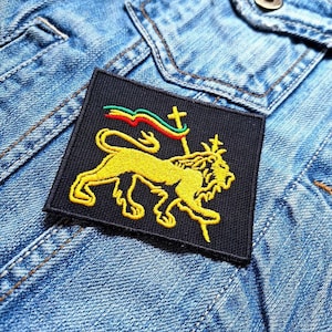 Puede incluir: Un parche negro con un león amarillo, una bandera roja, verde y amarilla, y una cruz. El león está de pie sobre sus patas traseras y tiene una corona en la cabeza.